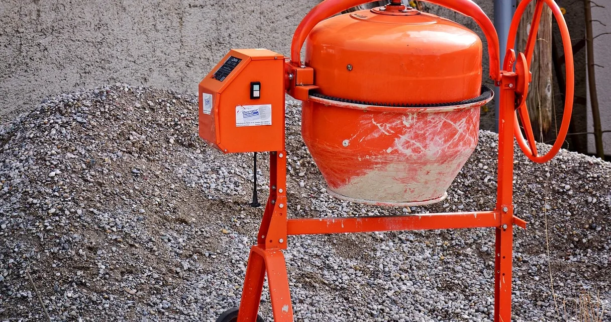 Betoneira laranja de obra ao lado de um monte de brita, com o tambor sujo de concreto