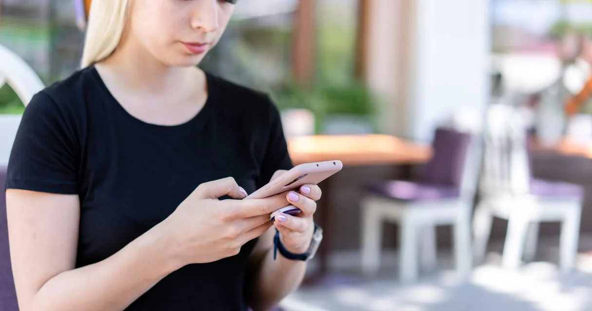Mulher de camiseta preta digitando no celular na área externa de um café